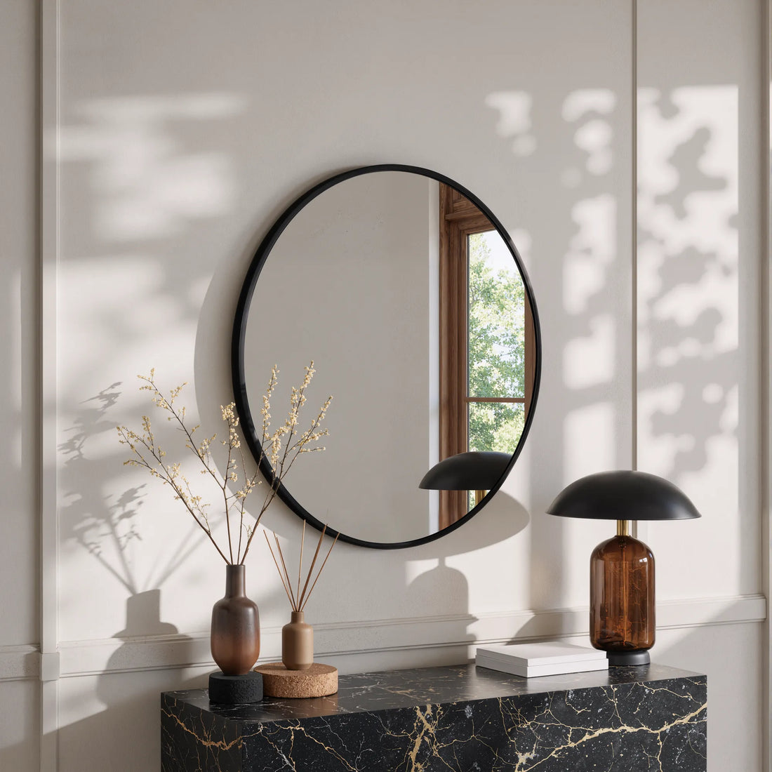 Sylvie black round wall mirror above a dark marble console with a dome table lamp and dried stems in a sunlit room